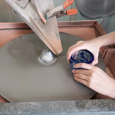 A person holds a glass bowl over a spinning lap wheel in a workshop.