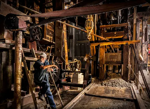 A man sits inside a rustic mill structure surrounded by large wooden and metal machinery.
