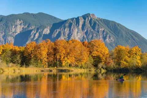 A person kayaking on a calm river with autumn trees and mountains in the background.