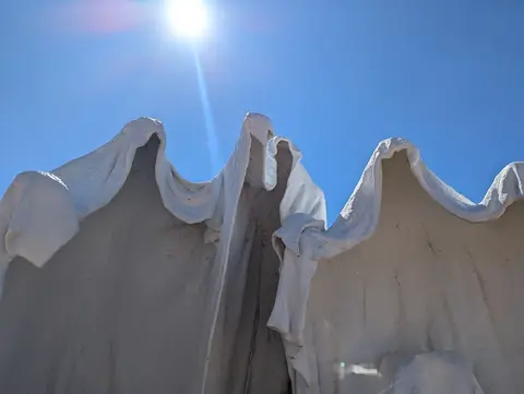White hooded statues stand against a bright blue sky with the sun shining above.