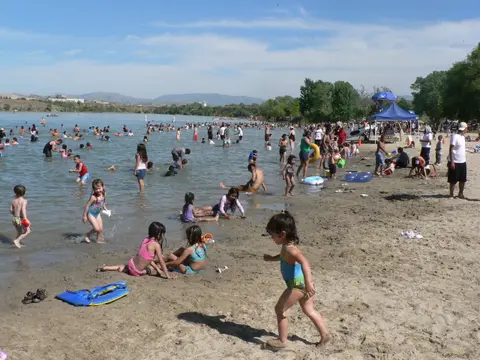 A crowded beach with people swimming and playing in the sand by a lake.