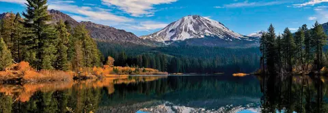 A scenic view of a snow-capped mountain reflecting in a calm lake with surrounding forests.