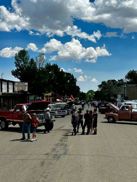 A street scene showing a classic car show with people gathered around vehicles under a cloudy blue sky.