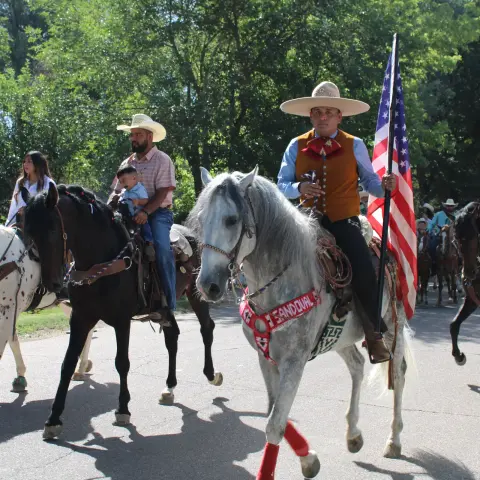 A group of people riding horses in a parade, with one person carrying an American flag.