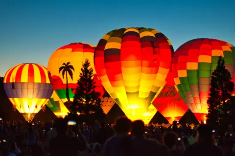 A group of illuminated hot air balloons glowing against the evening sky with spectators gathered below.
