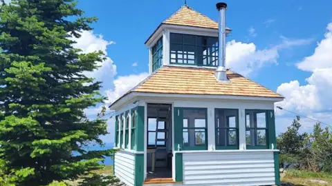 A small white and green lookout tower stands outdoors next to a large pine tree under a partly cloudy sky.