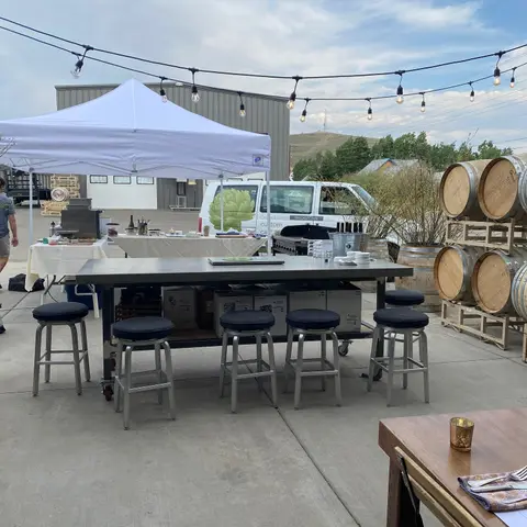 An outdoor event setup with tables, stools, a white canopy tent, and wine barrels.