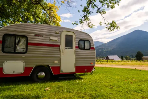 A vintage-style travel trailer parked in a grassy field with mountains in the background.