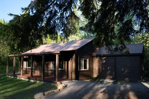 A dark wooden cabin with a metal roof and a covered porch stands among trees.
