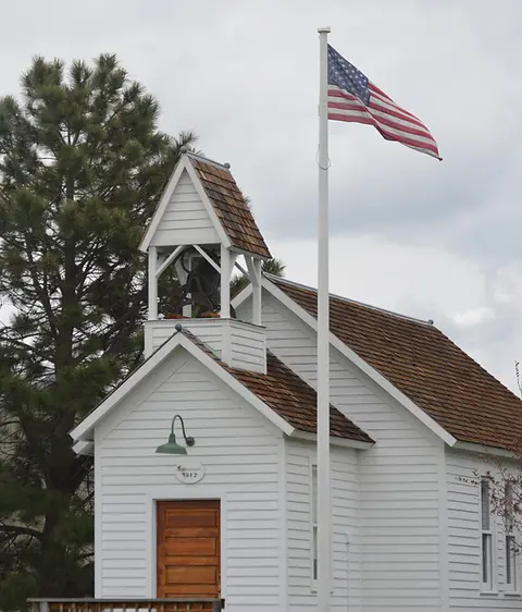 A small white wooden building features a bell tower and an American flag flying on a pole next to it.