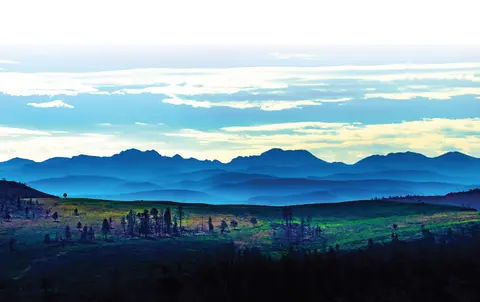A landscape view showing grassy hills with sparse trees and distant blue mountains under a cloudy sky.
