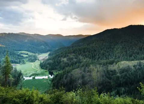 A scenic landscape view of a lush green valley surrounded by forested mountains under a cloudy sky.