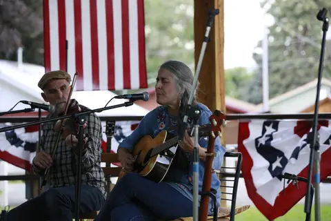 Two musicians perform outdoors with American flags hanging in the background.