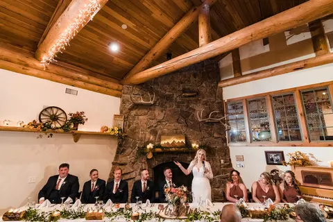 A bride speaking at a wedding reception table inside a rustic venue with a stone fireplace and wooden beams.