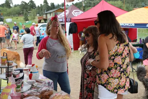 Three women converse near a vendor booth at an outdoor market.