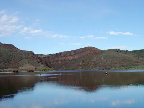 A scenic view of a calm lake surrounded by rolling hills and rocky cliffs under a blue sky.