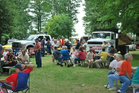 A group of people gathered outdoors near picnic tables and trucks.