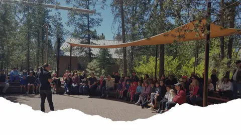 A group of people sitting on benches listening to a speaker outdoors.