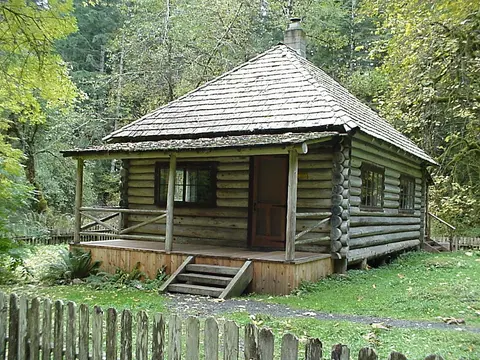 A log cabin with a porch stands among trees behind a wooden fence.