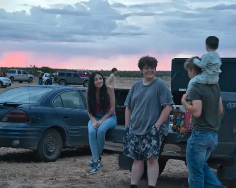 People gather around parked cars and trucks under a cloudy evening sky.