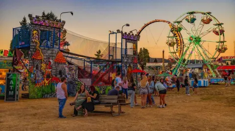 People enjoy a carnival at dusk with rides and a Ferris wheel.