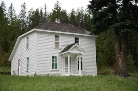A two-story white historic house stands among trees in a grassy clearing.