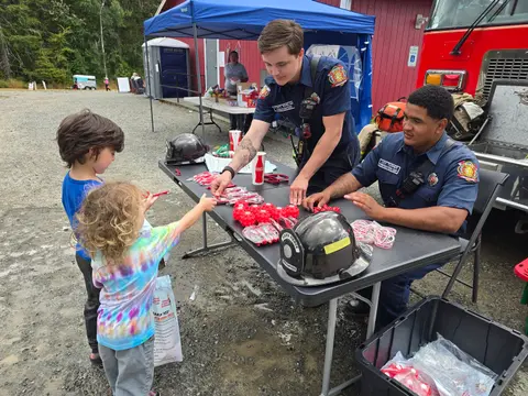 Two firefighters give items to children at an outdoor community event table.