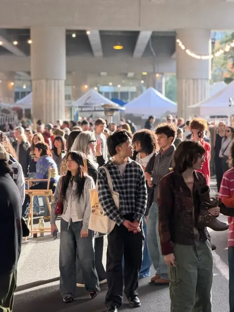 A crowd of people gathers under an overpass near market tents.