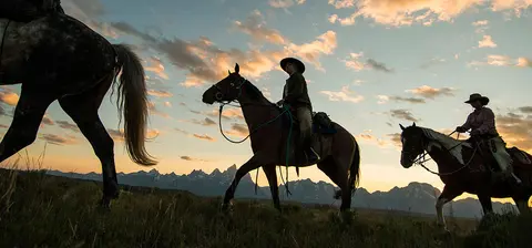 People horseback riding against a mountain backdrop at sunset.