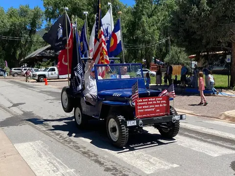 A blue jeep decorated with flags participates in a parade.