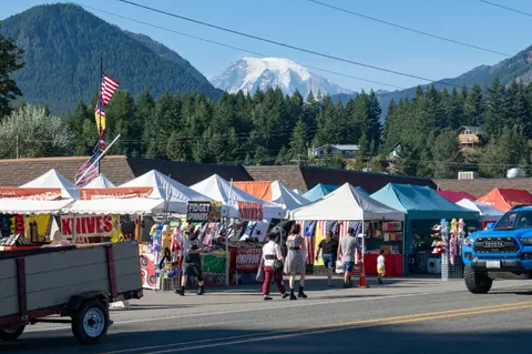 People walking along outdoor market stalls with a snowy mountain in the background.
