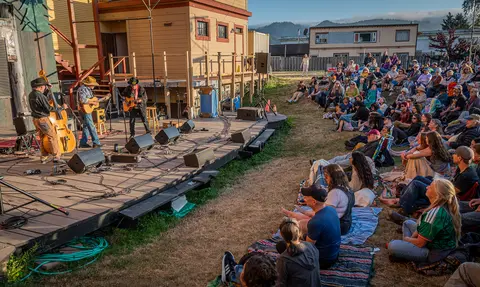 An outdoor band performs on a stage for a crowd seated on the lawn.