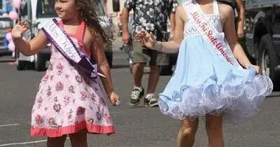 Two young girls wearing pageant sashes walk down a street during an outdoor event.