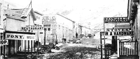 A historic black and white photograph shows a dirt street lined with old wooden buildings and business signs.