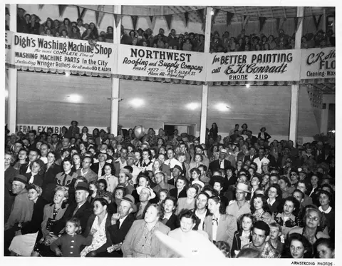 A black and white photograph showing a large crowd of people seated inside an event space with business signs hanging above.