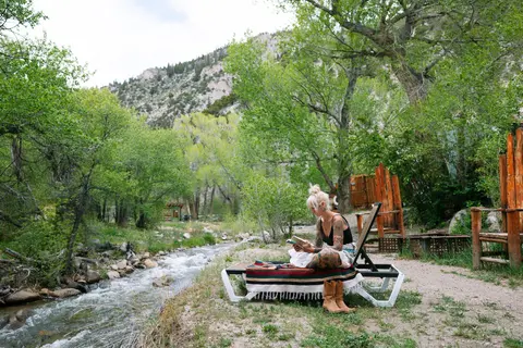 A woman reads a book on a lounge chair next to a flowing stream surrounded by trees and mountains.