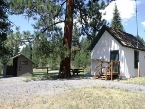 An exterior view of a small white building and an outhouse in a wooded area.