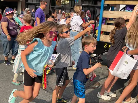Children run and reach for items near a parade float on a sunny street.