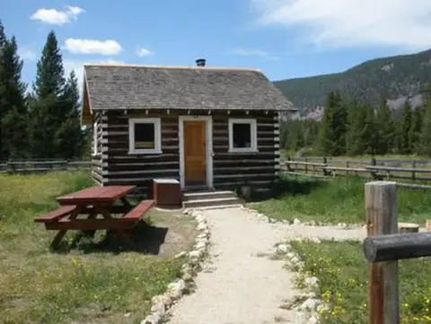 A small log cabin with a picnic table sits in a grassy clearing with mountains in the background.