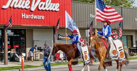 Two people ride horses carrying flags in front of a hardware store.