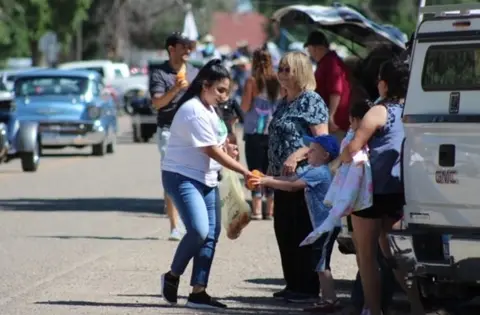 A woman hands food to a child on a street lined with cars.