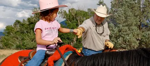 A child wearing a pink hat rides a horse alongside a man wearing a cowboy hat.
