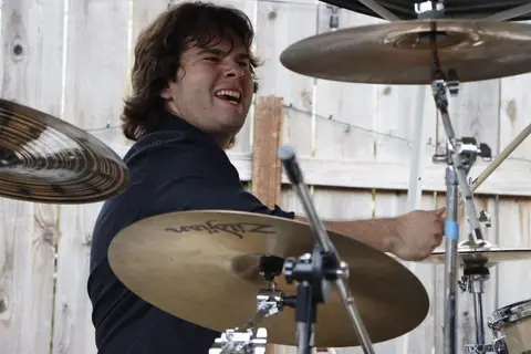 A smiling drummer plays his drum kit outdoors.