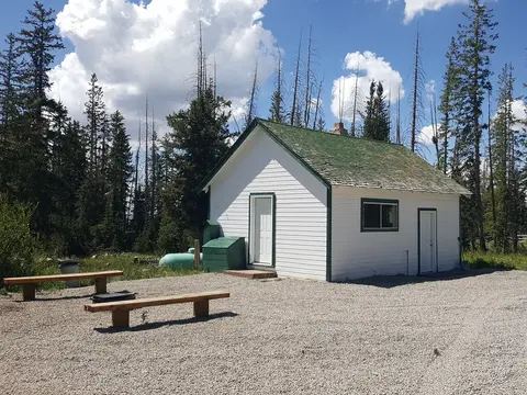 A small white building with a green roof sits in a clearing surrounded by trees.