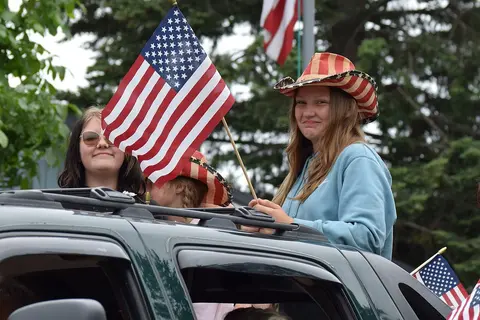 A young woman wearing a patriotic cowboy hat holds an American flag while riding in a vehicle.