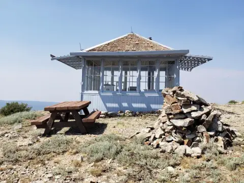 A small wooden lookout building sits on a hilltop with a picnic table and a rock cairn.