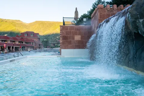 A waterfall flows into an outdoor swimming pool with buildings and mountains in the background.