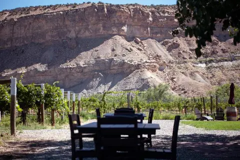 An outdoor dining area with tables and chairs in front of a vineyard and large rocky cliff.