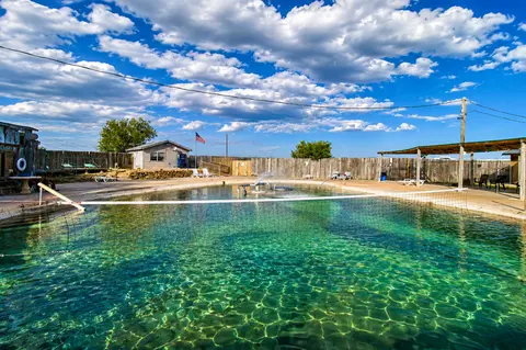 An outdoor swimming pool with a volleyball net under a blue sky with clouds.