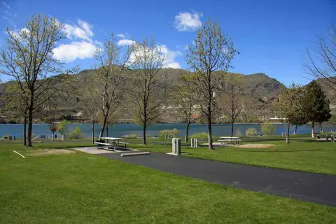 A grassy park with picnic tables alongside a body of water and mountains in the background.